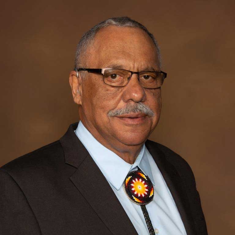 Portrait of Greg Jacobs, wearing a brown suit, white shirt, and beaded bolo tie.